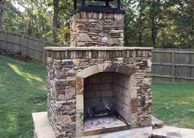 Outdoor stacked stone fireplace with black cover and wood fence in the background